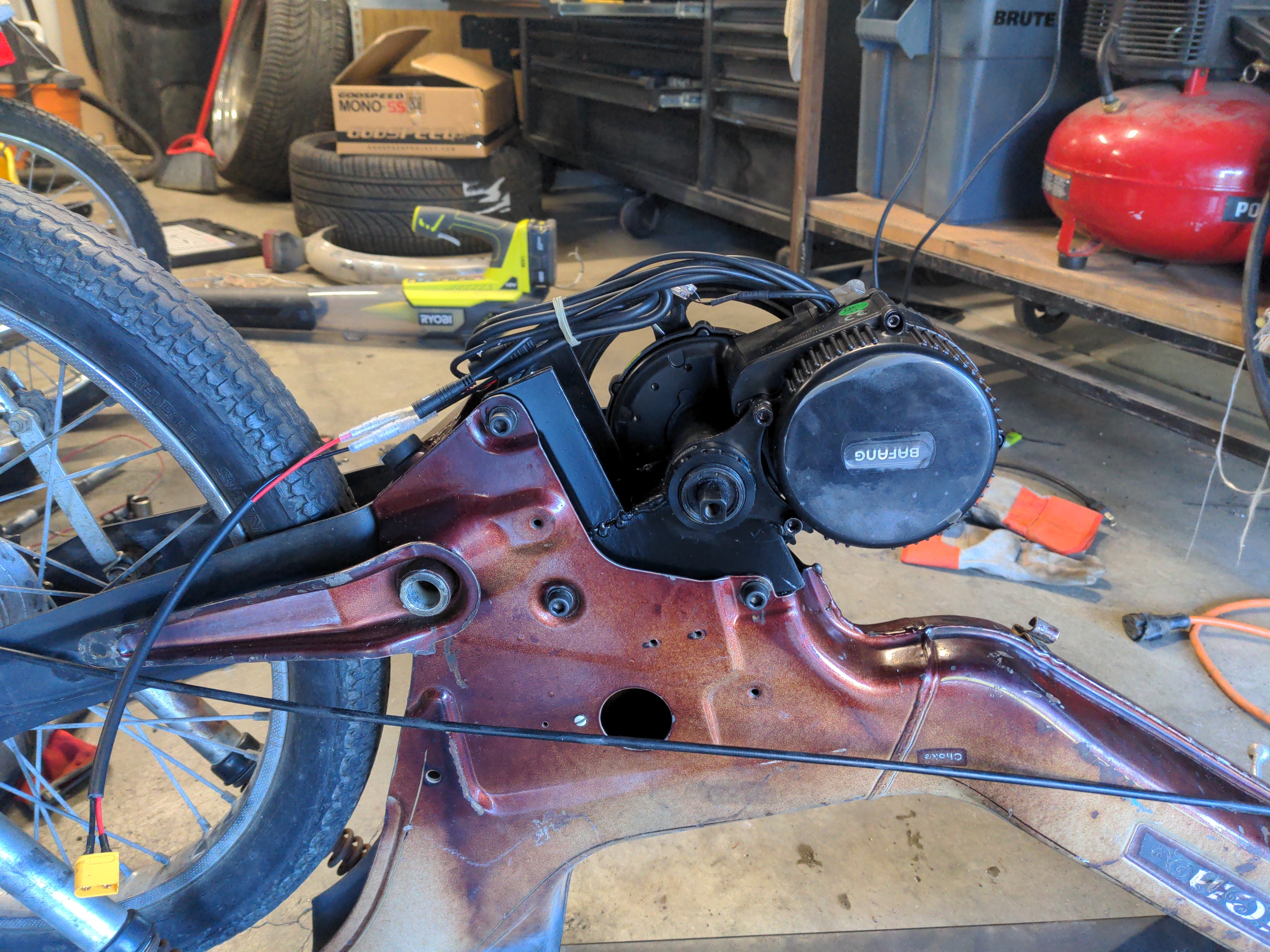 A custom fabricated metal bracket, slotted into the frame of a Puch moped. The electric motor has been mounted to the bracket, and is locked into position. The bracket has been painted black.