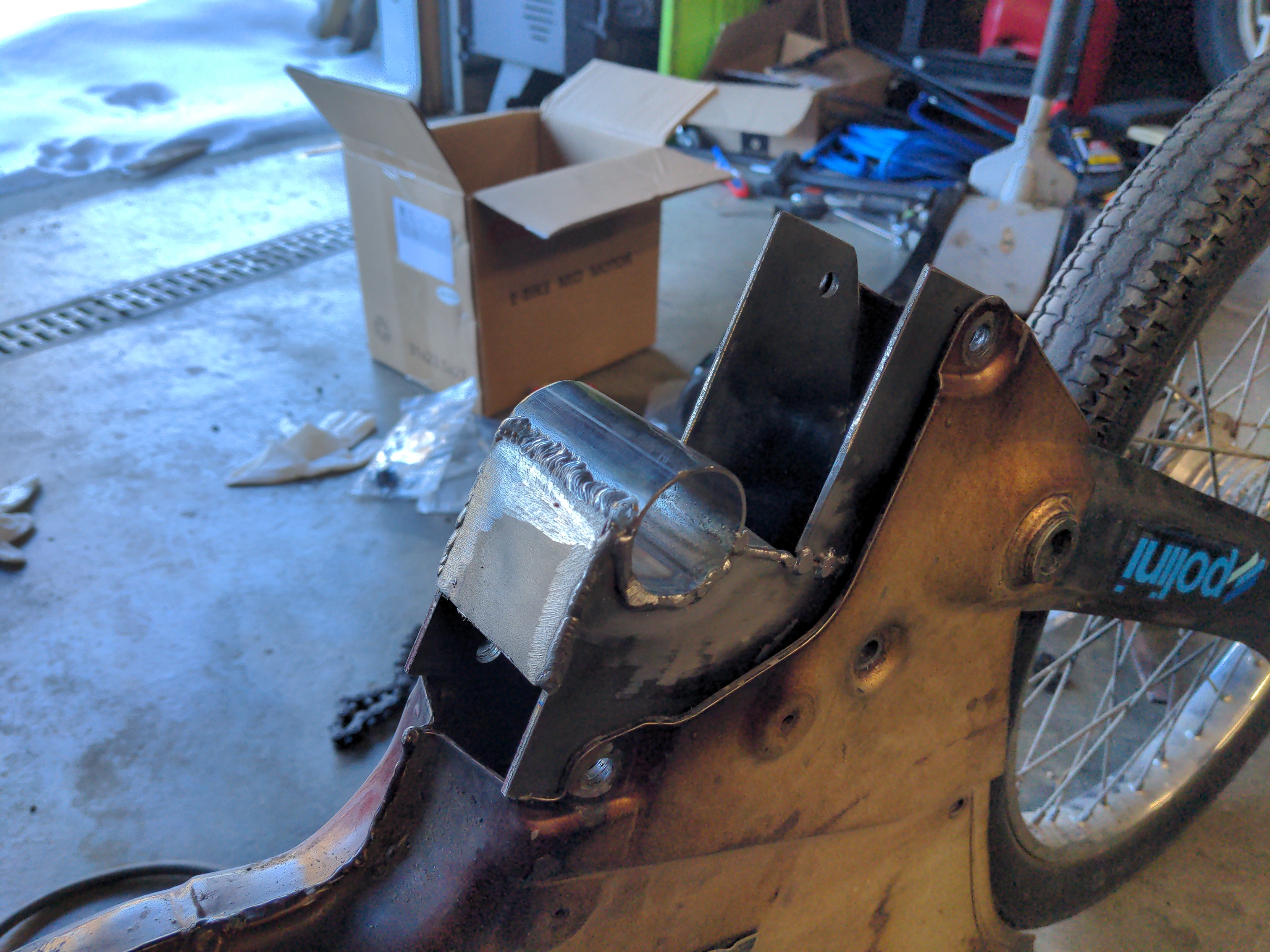 A custom fabricated metal bracket, slotted into the frame of a Puch moped. The bolt holes in the bracket line up nicely with the holes in the frame. The welds are a little rough, but serviceable.