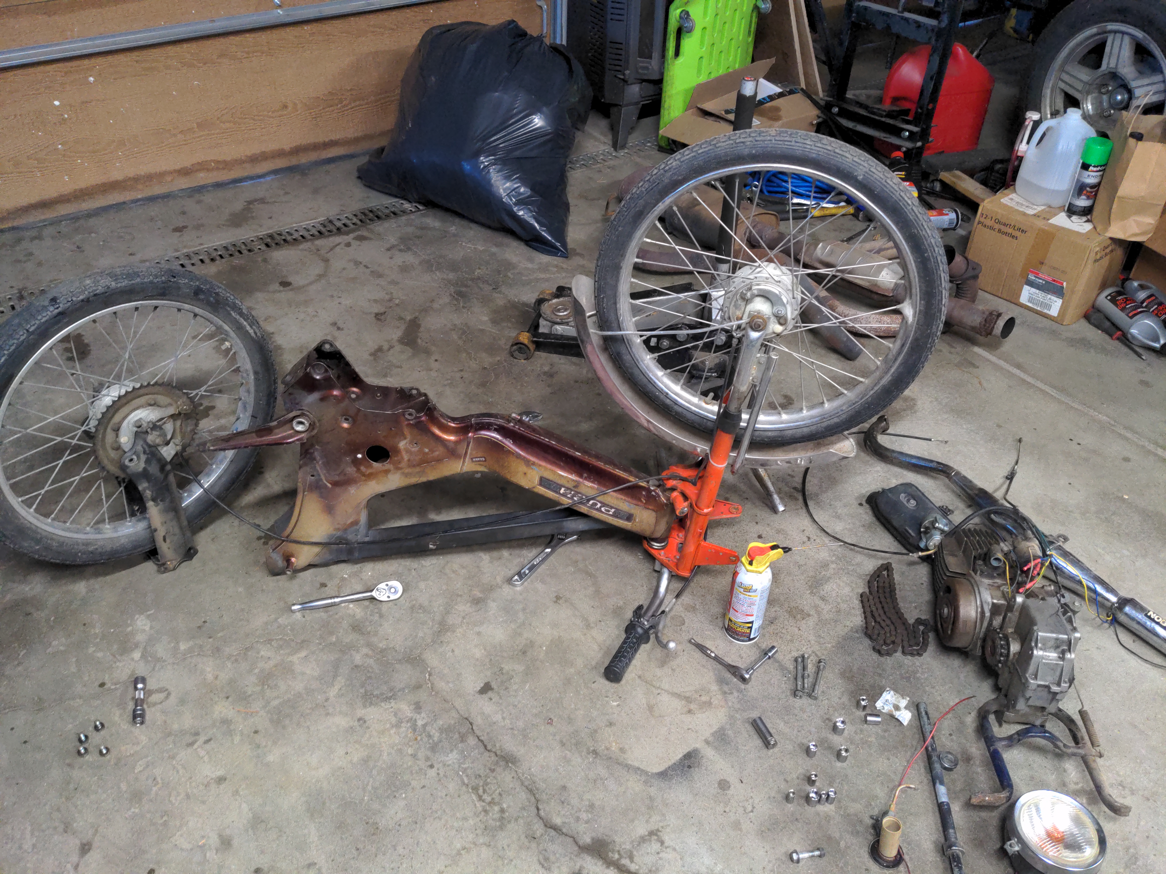 A partially disassembled Puch moped. The moped is resting upside down on a concrete floor. The front fork and wheel are still connected, but everything else has been uninstalled. The rear suspension and wheel are resting to the left of the body. To the right, a pile of discarded parts illustrates just how much weight was removed for this project.