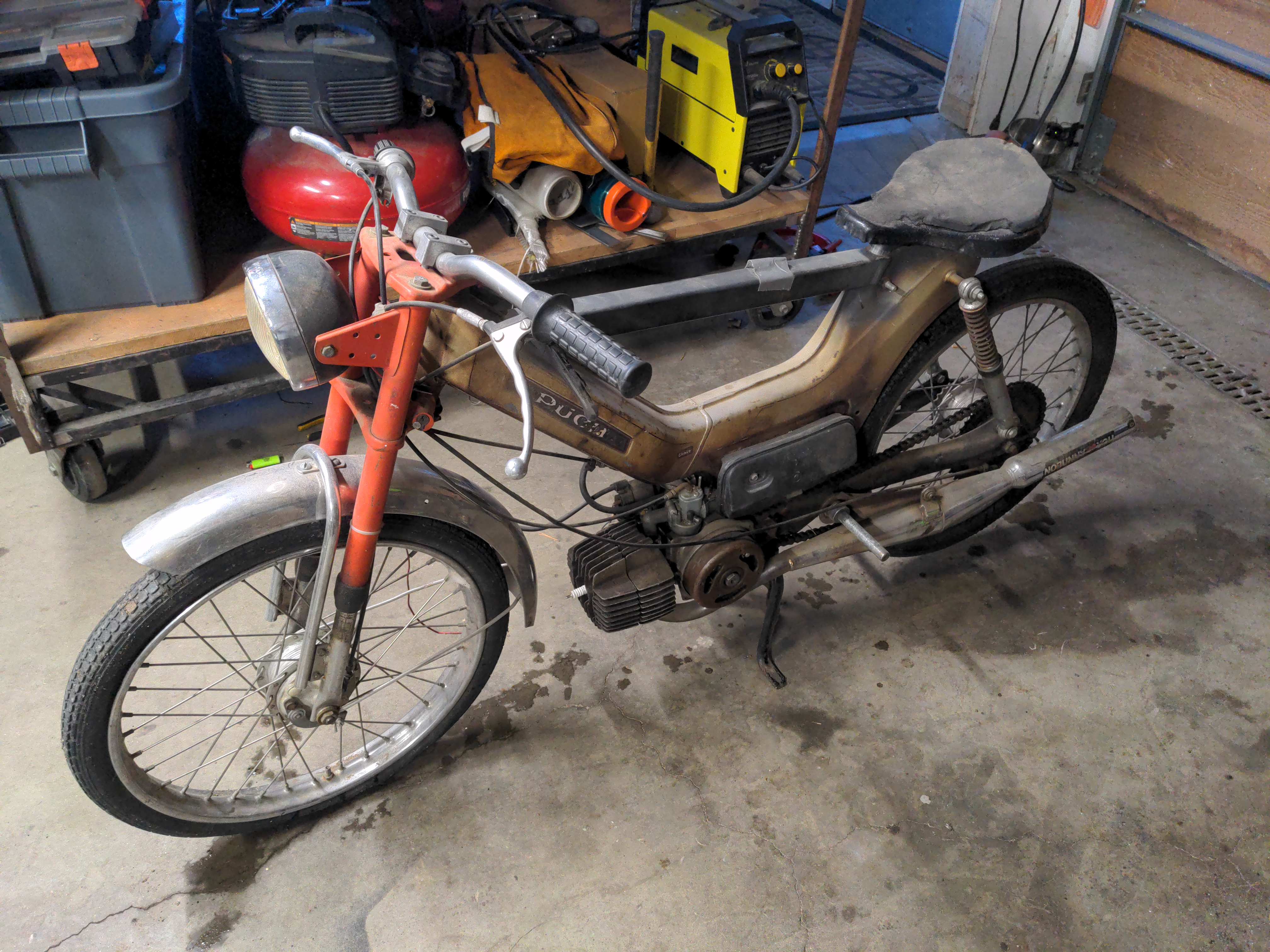 An old Puch moped sitting in a garage. The moped has already been modified with a long bar that runs front to back (presumably to attach equipment or add structural stability). The moped is clearly worn out, and lots of blemishes, rust, and grease spots are visible on what's left of the paint.