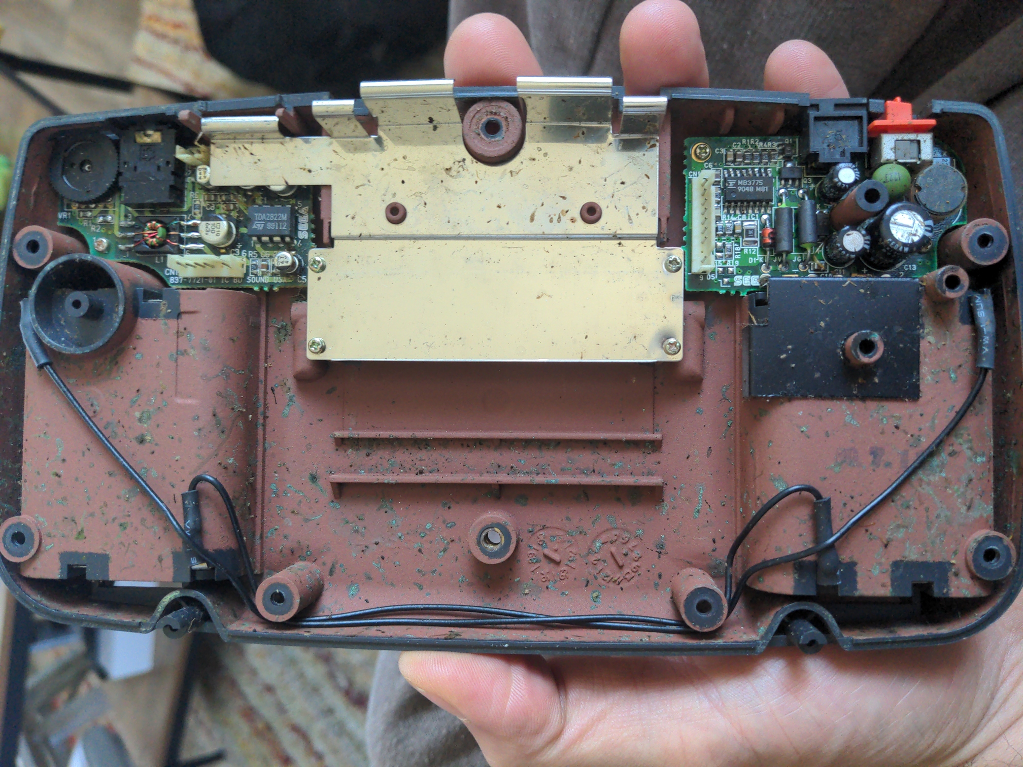 The outer shell of a Sega Game Gear, rotatedto show the inside. The main board has been removed, but the original audio and power boards are still present. Also present is a gross collection of tiny bug parts and related stains.